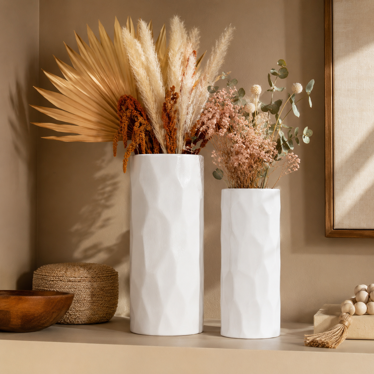 Two white textured ceramic vases filled with dried grasses and eucalyptus stems, styled on a beige shelf with a wooden bowl and woven decor in a warm neutral room.