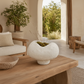 A textured off-white stone planter with a wavy, organic shape sits on a rustic wooden coffee table in a bright, modern boho-style living room. The planter is filled with green moss and dried grass stems, set against a warm, neutral interior with a sofa, potted plant, and an open doorway leading to a garden view.