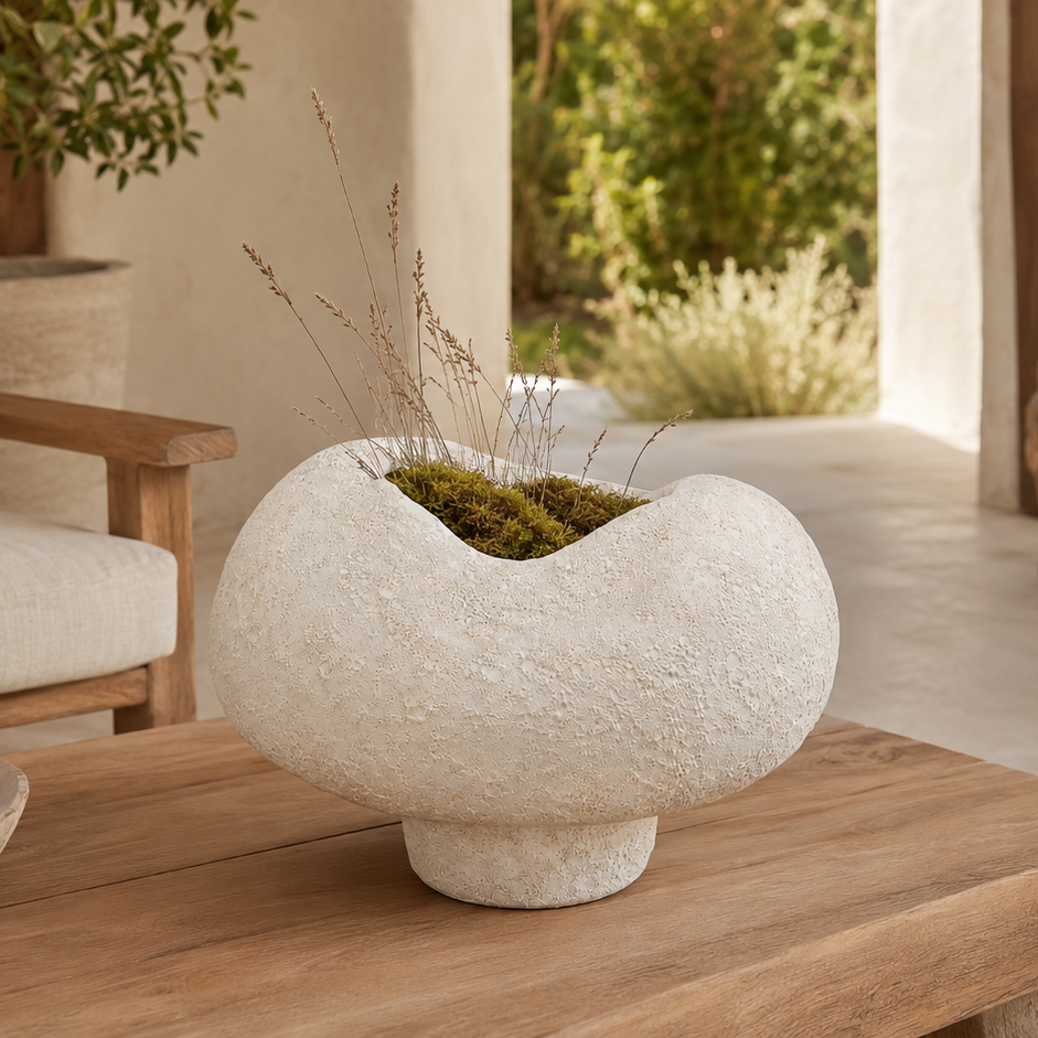 A textured, organic-shaped off-white stone planter vase filled with green moss and dried grasses, displayed on a wooden coffee table in a modern, sunlit living room.