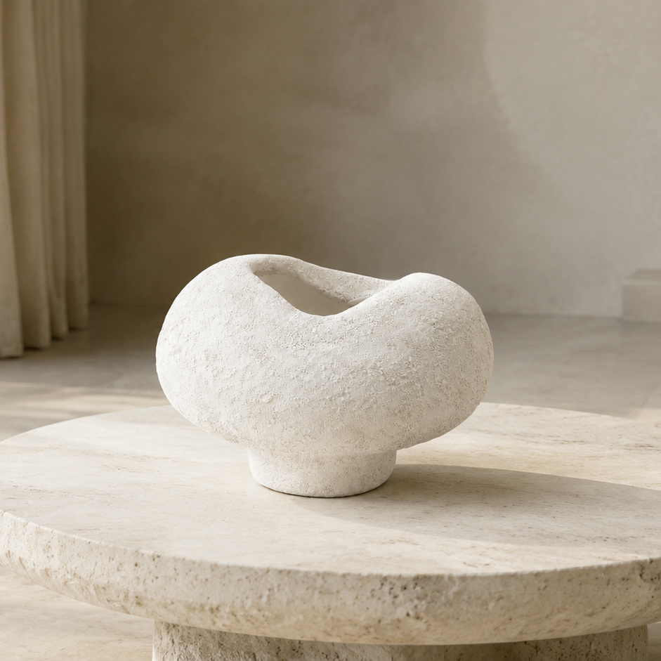 A sculptural, organic-shaped white stone vase displayed on a round stone coffee table in a minimalist, warm-toned modern living room.