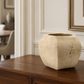 A textured beige stone planter pot with a hexagonal shape, displayed on a dark wood sideboard in a beautifully decorated room with white walls and framed art.