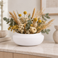 A white ceramic bowl vase filled with a dried floral arrangement featuring golden wheat stalks, yellow globe flowers, white daisies, and green eucalyptus leaves, displayed on a light marble countertop.