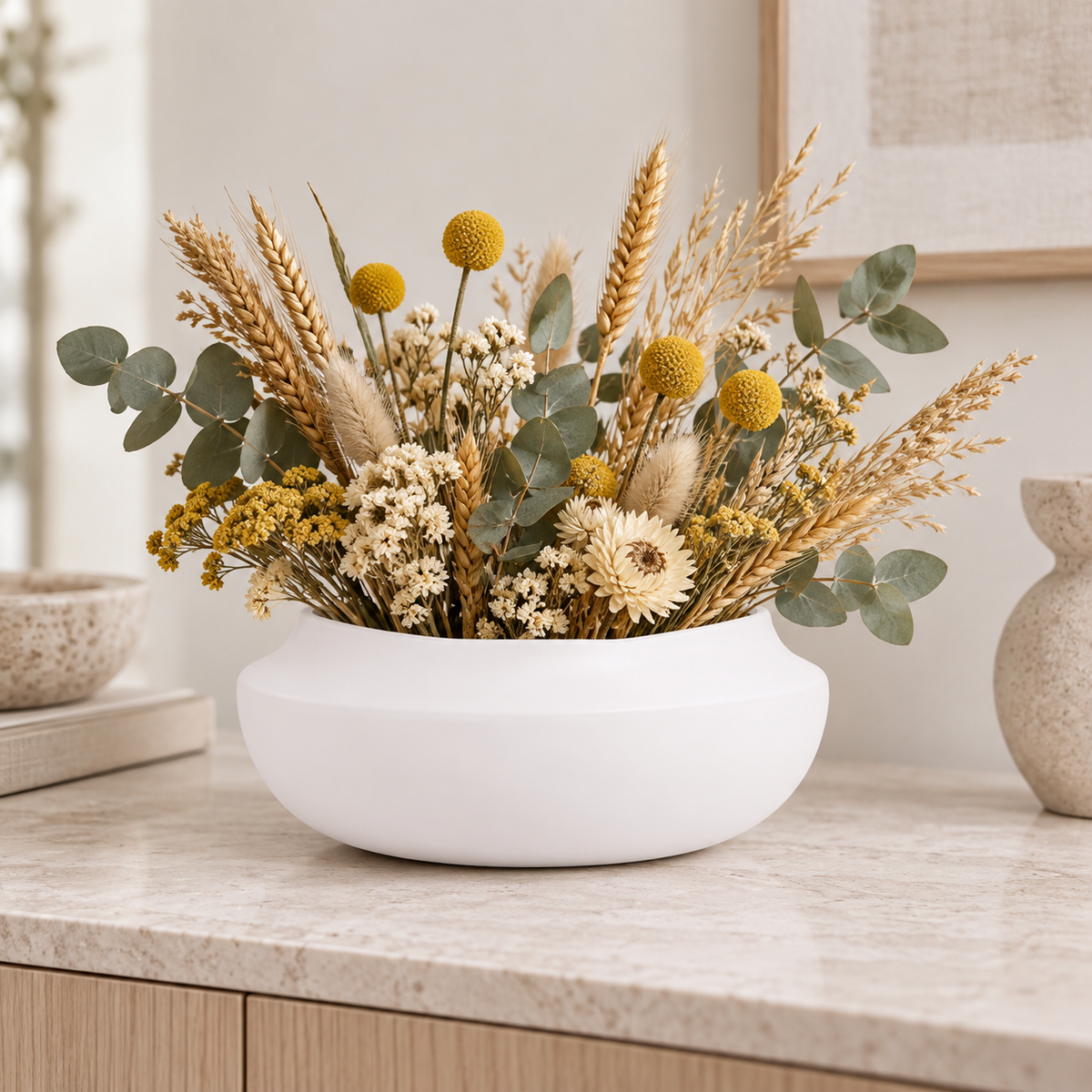 A white ceramic bowl vase filled with a dried floral arrangement featuring golden wheat stalks, yellow globe flowers, white daisies, and green eucalyptus leaves, displayed on a light marble countertop.