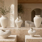 A set of sculptural, organic-shaped off-white ceramic vases displayed on stone plinths inside a modern, sunlit room with beige walls and large windows.