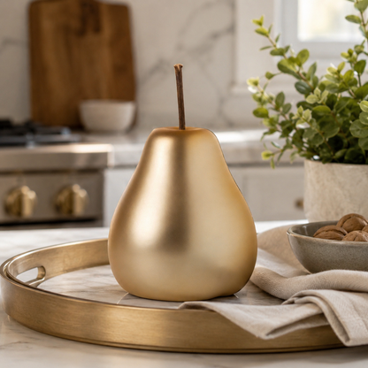 A metallic gold decorative pear-shaped object with a short stem stands on a round gold tray draped with a beige cloth in a bright kitchen setting.
