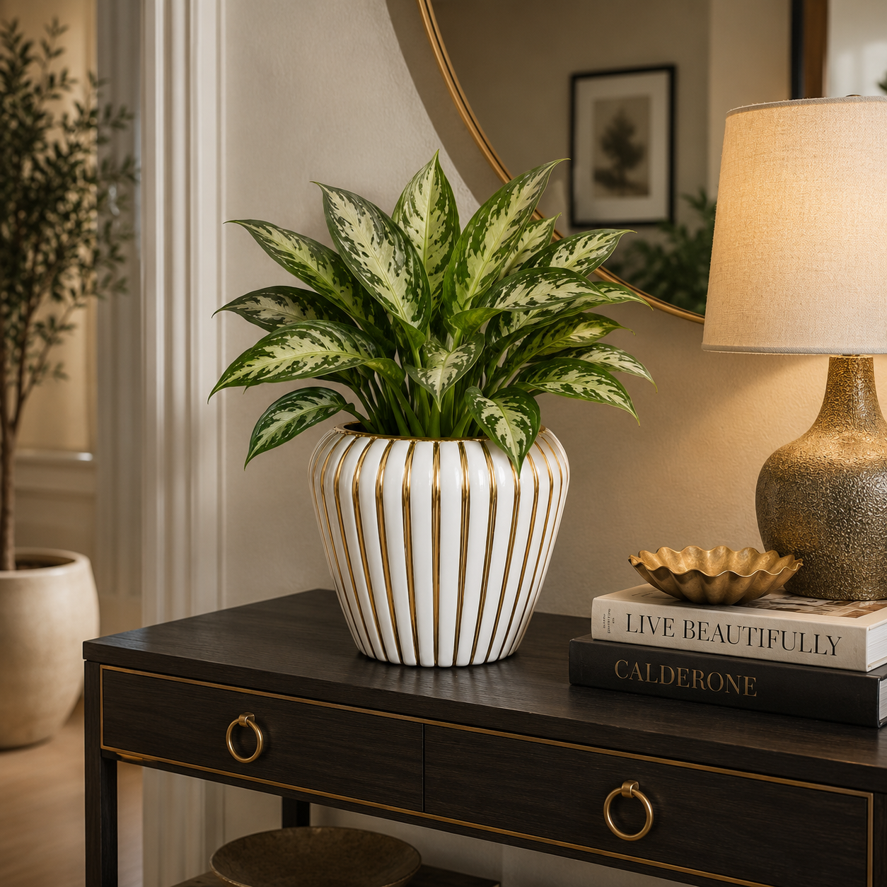 A potted houseplant with green and white variegated leaves sits on a dark wood console table in a styled interior. The plant is in a white ribbed ceramic pot with vertical gold-toned stripes.