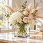A floral arrangement in a clear square glass vase on a marble countertop, featuring white hydrangeas, blush pink roses, pampas grass, and green foliage in a bright indoor setting.