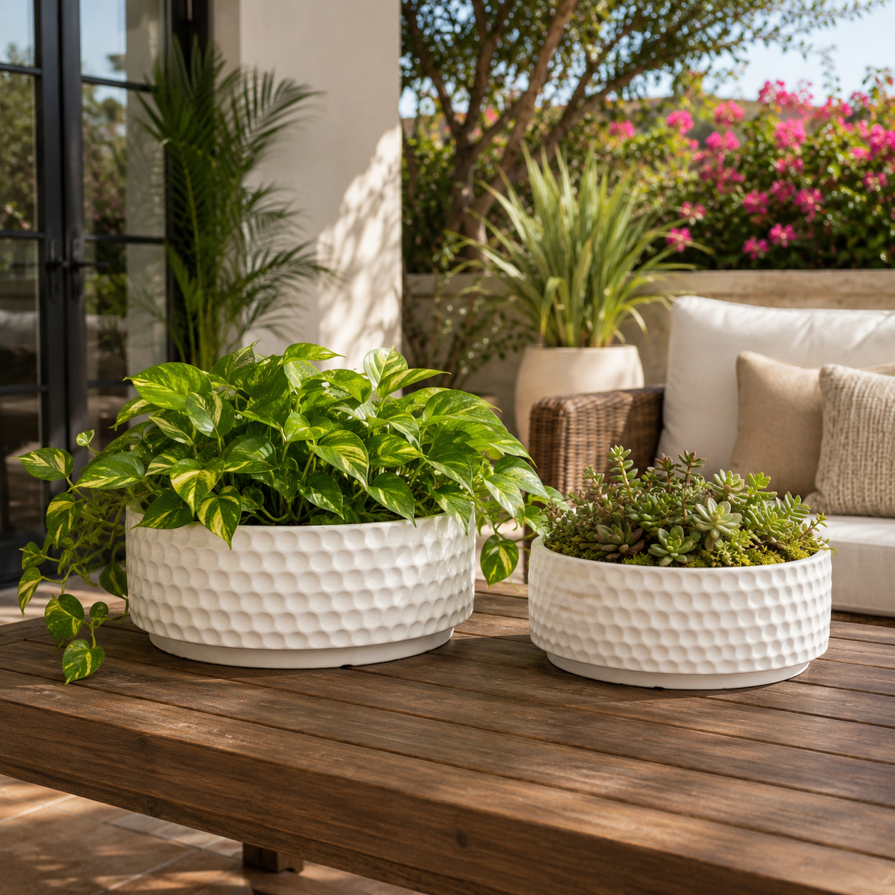 Two white textured planter pots with green plants displayed on a wooden outdoor patio table next to a white sofa.