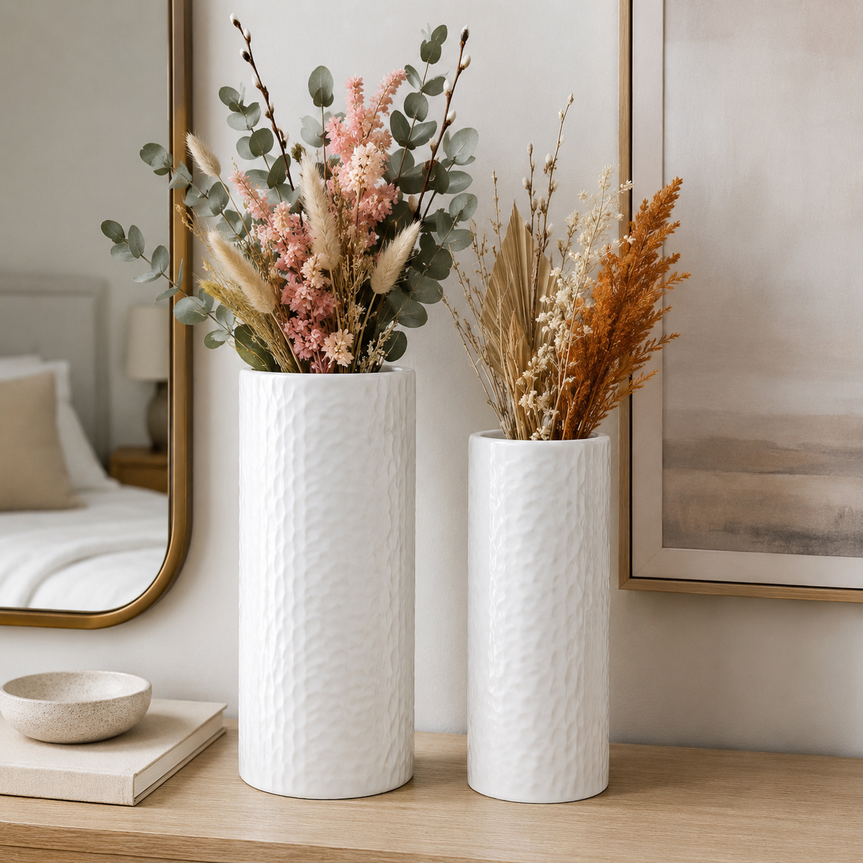Two tall textured white ceramic vases filled with dried floral arrangements, displayed on a wooden console table in a softly styled room with a mirror and framed wall art in the background.