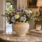 A large cream-colored ribbed ceramic urn vase filled with a lush floral arrangement of purple, blue, white, and green flowers, placed on a marble-topped table in a softly lit indoor setting.