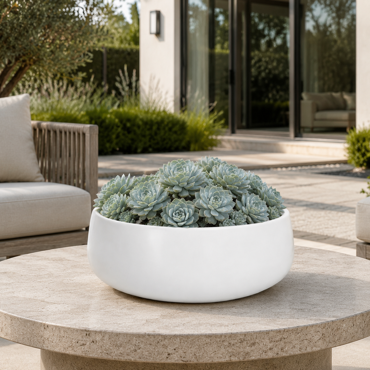 A round white ceramic planter filled with pale blue-green succulents, displayed on a stone patio table in front of a modern outdoor living area.